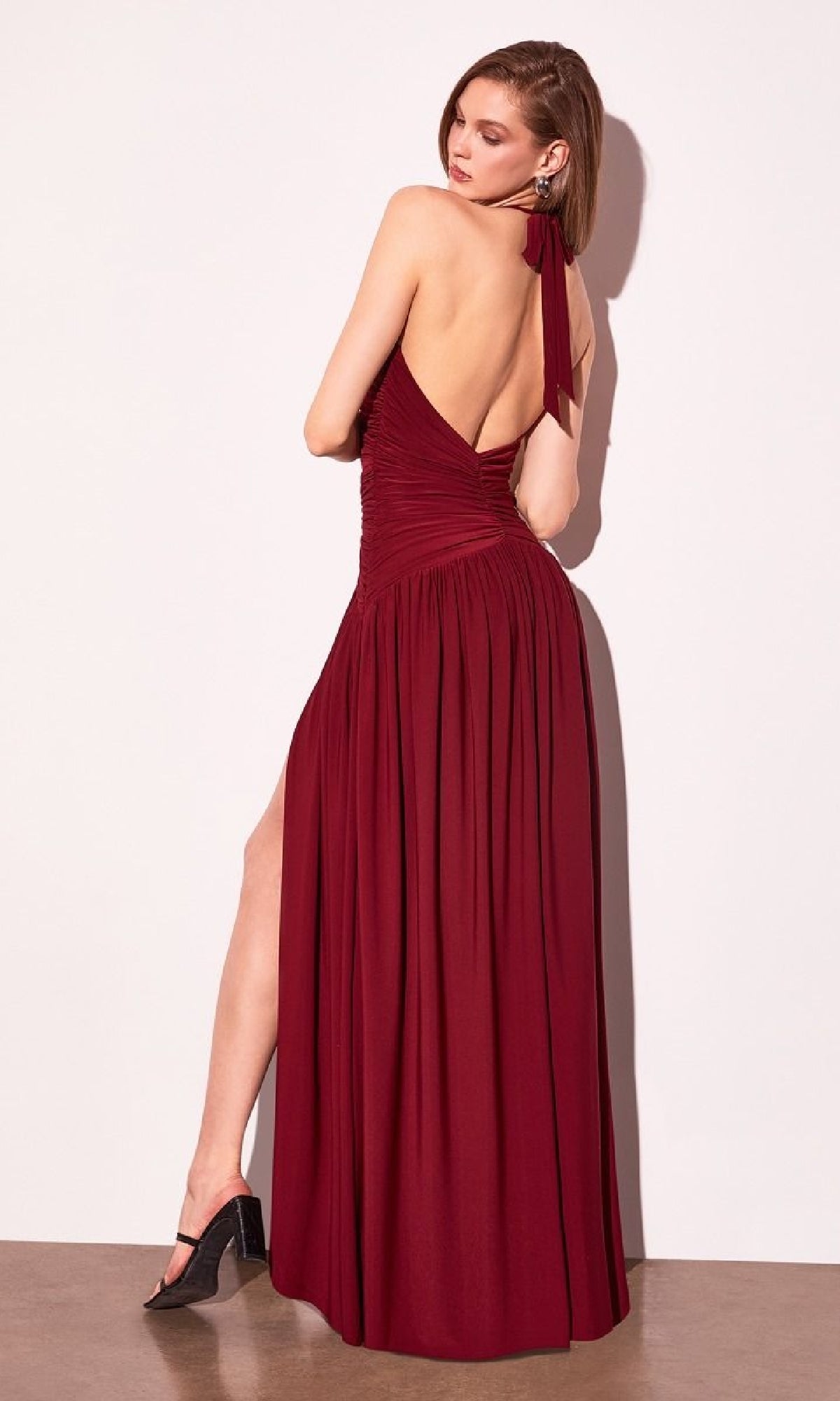 A woman in the Amoris T502 Backless Halter Prom Dress with Slit by Cinderella Divine, a sleeveless burgundy gown, stands against a white wall and looks over her shoulder.